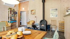 Een knus woongedeelte met houten details, een open haard, een eettafel met brood en boter, en een boekenkast.