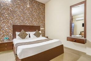 A neatly made bed with a wooden headboard, a bedside table with a bottle, and a mirror above a dresser. - iROOMZ Hotel Royal Hill Park (Hyderabad)
