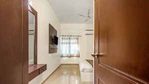 A bedroom with a wooden door, a ceiling fan, a bed, a TV mounted on the wall, and a window with curtains. - iROOMZ Hotel Royal Hill Park (Hyderabad)