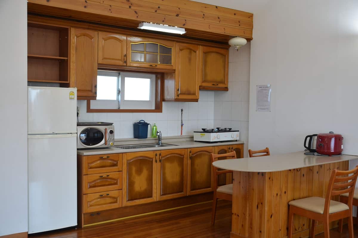 Een keuken met houten kastjes, een koelkast, een magnetron, een spoelbak en een fornuis. Er is een eettafel met stoelen.