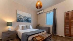 A bedroom with a bed, bedside tables, a nightstand, a wall-mounted air conditioner, a window with curtains, and a wooden wardrobe. - La Mona Suites 2 Bedroom Family Apartment (San Juan)