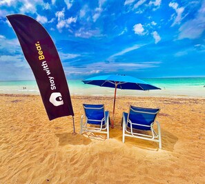 A beach with two blue beach chairs, a blue umbrella, and a black flag with text. - La Mona Suites 1 Bedroom Apartment with Rooftop Terrace (San Juan)