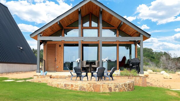 Una casa moderna con un amplio ventanal de vidrio, un patio de piedra y un área de comedor con mobiliario exterior.