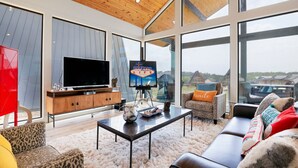 Una sala de estar moderna con un televisor grande, techo de madera y ventanas amplias con vistas del entorno.