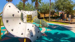 A playground with climbing structures and a slide, surrounded by palm trees and a fenced area. - New! Lesa's Disney Vacation Home (Davenport)