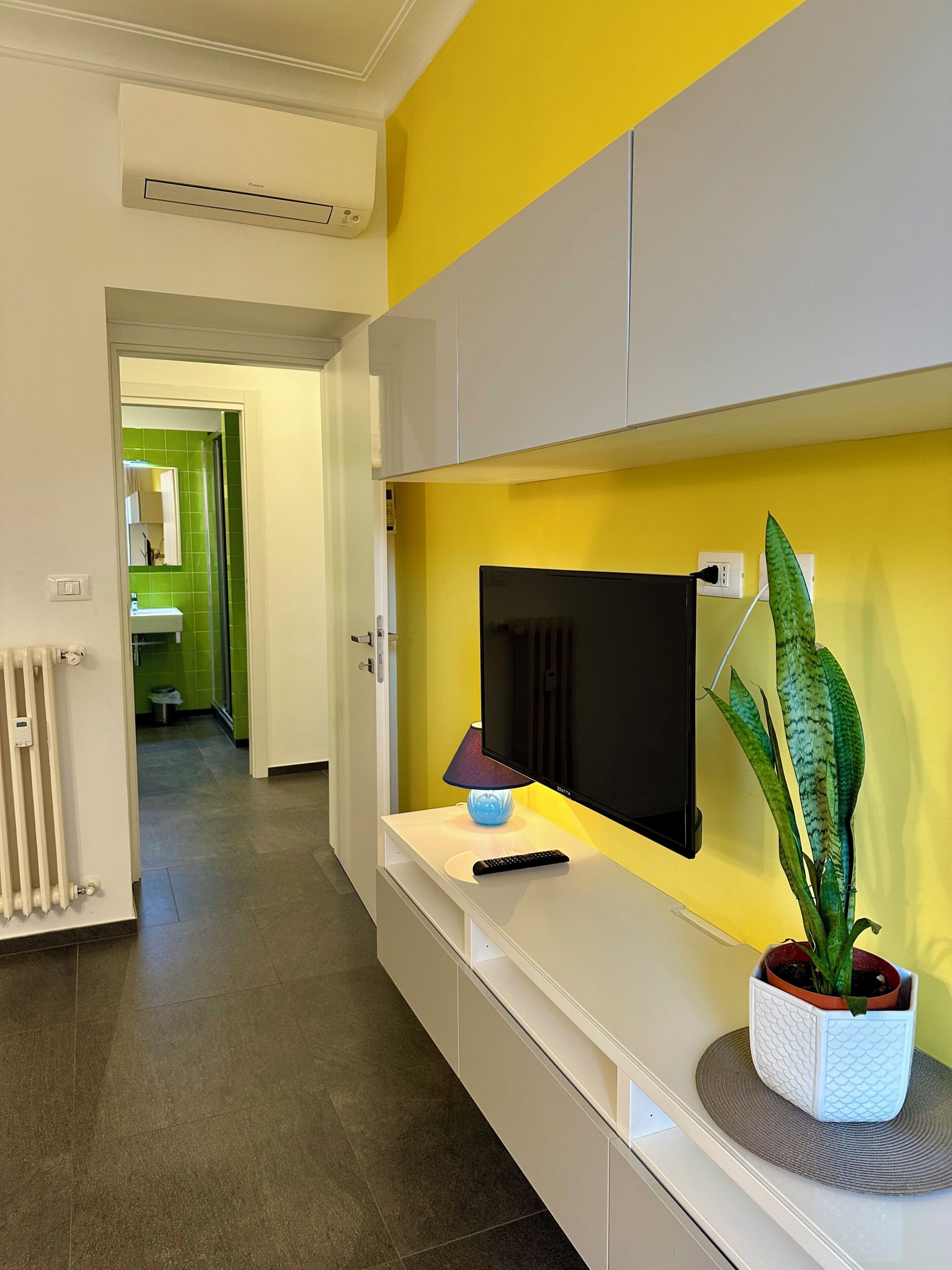 Ein Flur mit einem weißen Schrank, einem flachbildfernseher an einer gelben Wand, einer Fernbedienung und einem Topfpflanzen.