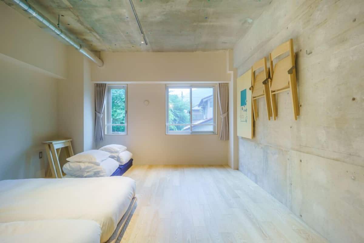 A room with a bed, two windows, a wooden wall, and a ceiling with exposed pipes.