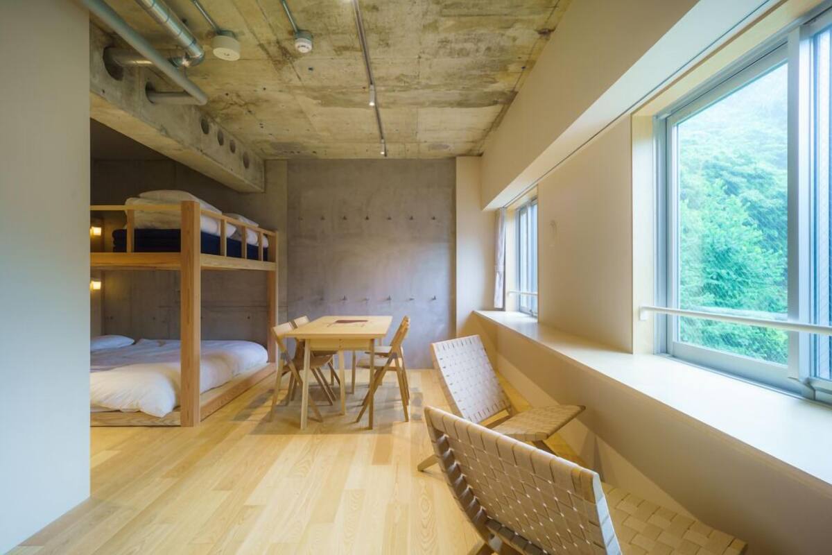 A room with bunk beds, a table, chairs, a window with a view, and exposed ceiling pipes.