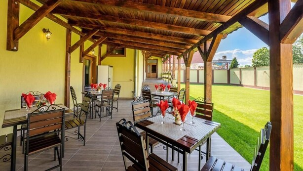 An outdoor patio with wooden tables and chairs, a covered area with a wooden ceiling, and a view of a green lawn and buildings in the background. - Hotel PrincesSophie (Sighisoara)