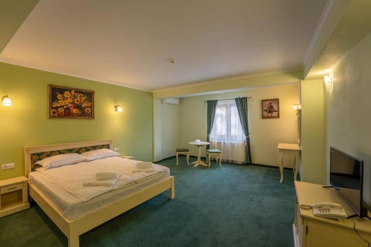 Habitación de hotel con cama, escritorio con teléfono y televisor, una mesita con una silla y una ventana con cortinas.