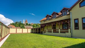A courtyard with a lawn, a building with a balcony, and a tower in the background. - Hotel PrincesSophie (Sighisoara)