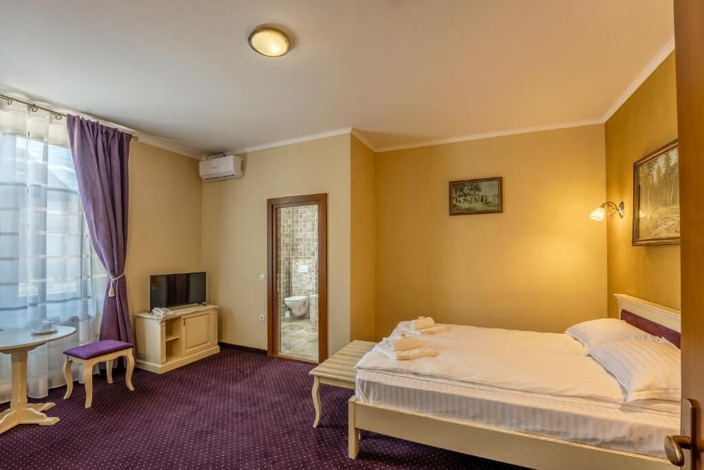 Habitación de hotel con cama, escritorio con televisión, una mesita y baño visible a través de una puerta de vidrio.