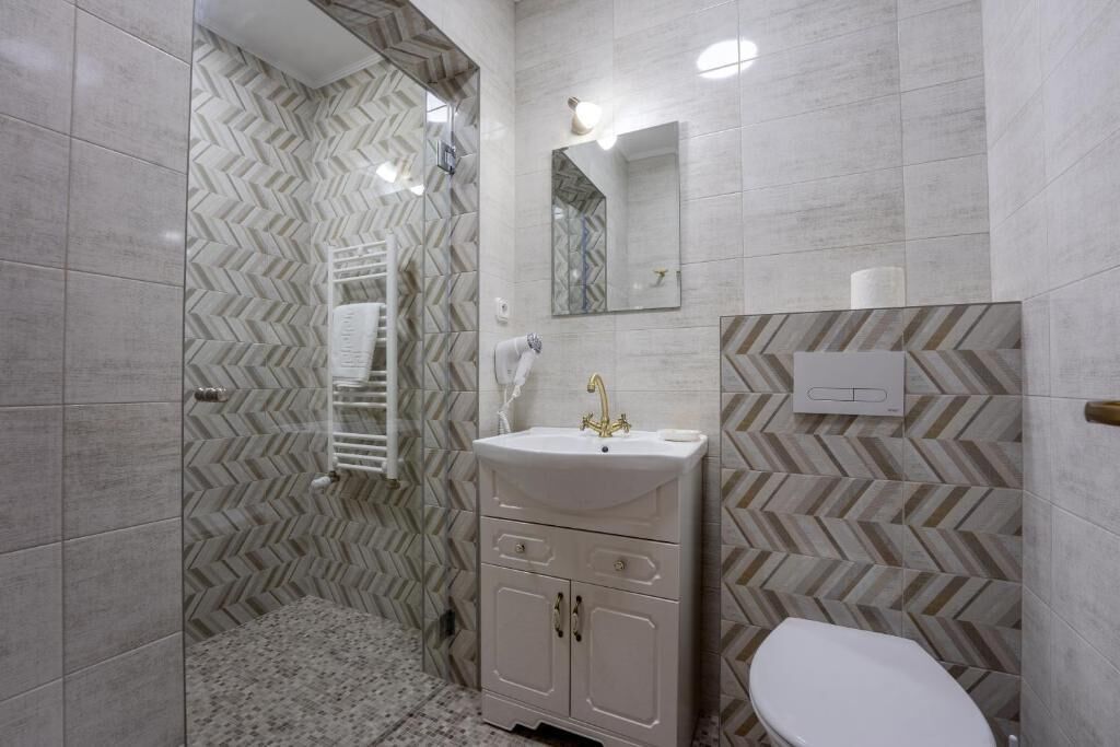 Un bagno con parete rivestita in piastrelle a motivo a spina di pesce, un lavandino bianco con rubinetto dorato, uno specchio sopra il lavandino e un water.