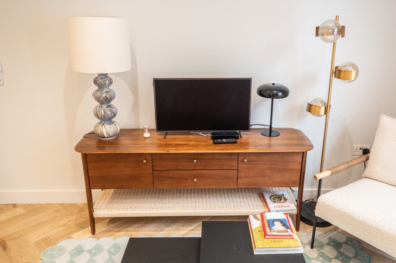Un escritorio de madera con un televisor, una lámpara y una lámpara de pie. Hay libros y un control remoto sobre el escritorio.