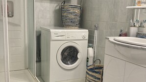 Un lavadero pequeño con una lavadora de carga frontal, un armario pequeño encima, una cesta tejida azul y blanca, y un tapete de madera en el suelo.