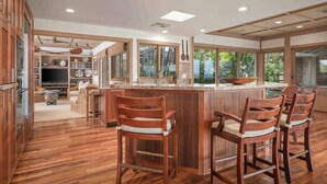 A spacious kitchen with wooden flooring, a central island with bar stools, and large windows offering views of greenery. - Fairways South 23 Four Bedroom (Kamuela)