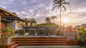A patio with outdoor seating, a stone wall, and a view of palm trees and a sunset. - Fairways South 23 Four Bedroom (Kamuela)
