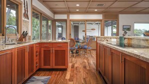 A modern kitchen with wooden cabinets, a granite countertop, and a dining area with wicker chairs. - Fairways South 23 Four Bedroom (Kamuela)