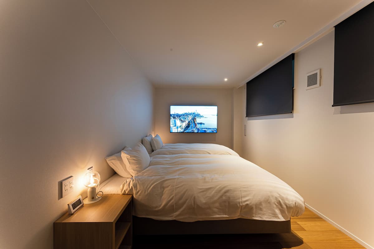 Ein modernes Schlafzimmer mit einem Bett, einem Nachttisch mit Lampe und einem an der Wand montierten Fernseher, der eine Stadtansicht zeigt.