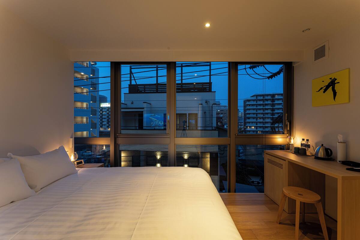 Ein modernes Hotelzimmer mit einem großen Bett, einem Schreibtisch mit Hocker und Blick auf die Stadt durch große Fenster.