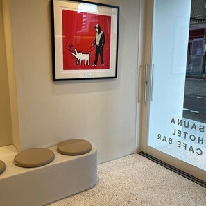Una obra de arte enmarco que muestra a una persona con un perro con correa, una zona de estar con bancos, y una puerta de cristal con texto.