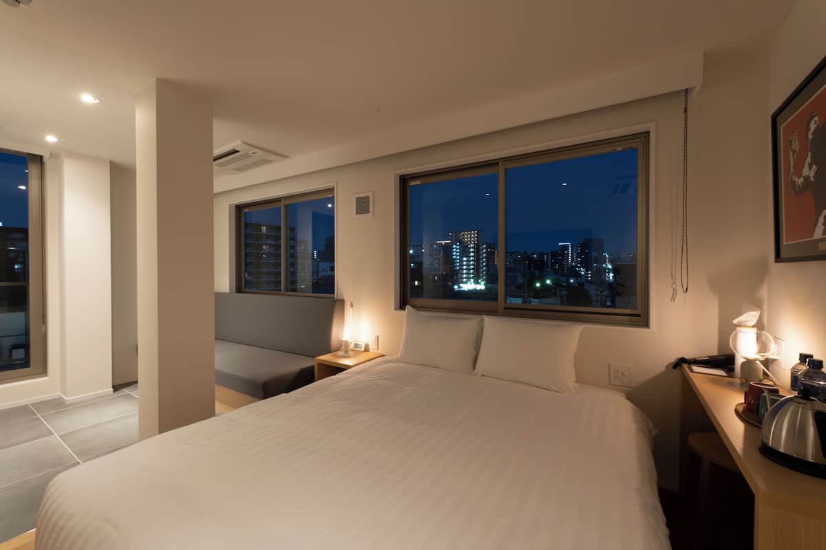 Ein Hotelzimmer mit einem großen Bett, einem Schreibtisch mit Lampe, einem Stuhl und Blick auf die nächtliche Skyline.