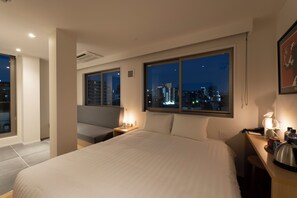 Habitación de hotel con una cama grande, un escritorio con lámpara, una silla y vista a la ciudad de noche.