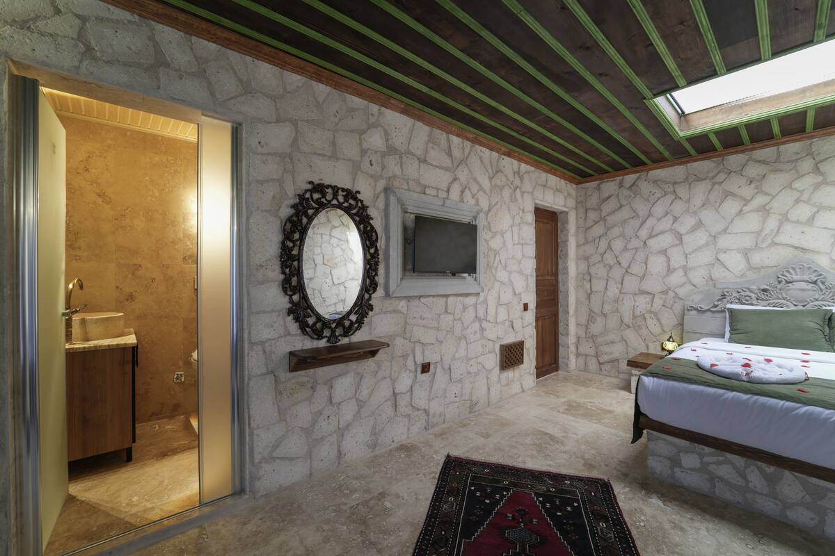 Quarto com parede de pedra, cama com colcha verde, televisão, espelho e armário de madeira.