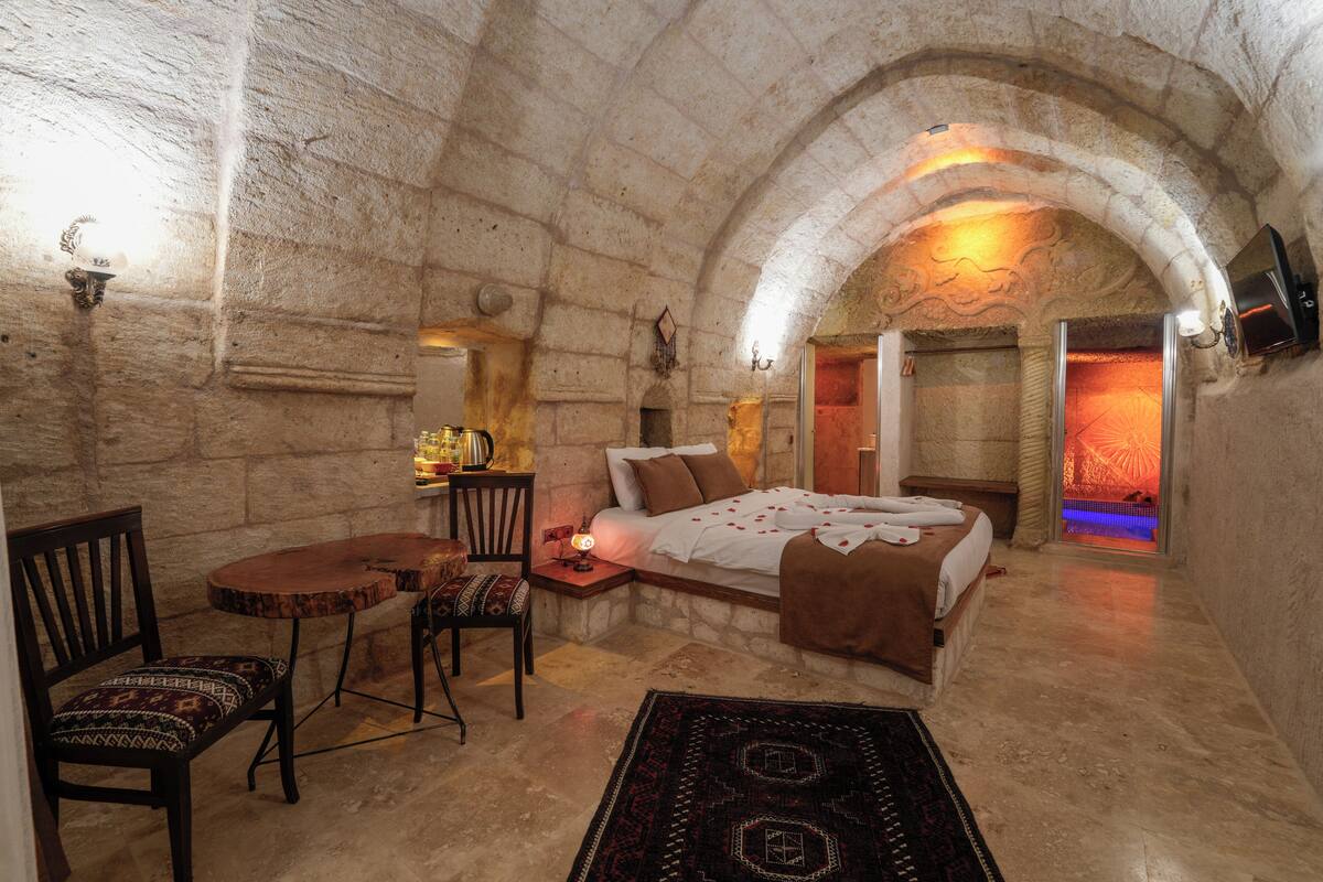 Quarto com paredes de pedra, uma cama, uma mesinha, uma cadeira e um tapete.