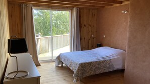 Ein Schlafzimmer mit Holzboden, einem Bett mit floralem Überwurf, einem Schreibtisch mit Lampe und einer Glastür zu einem Balkon.
