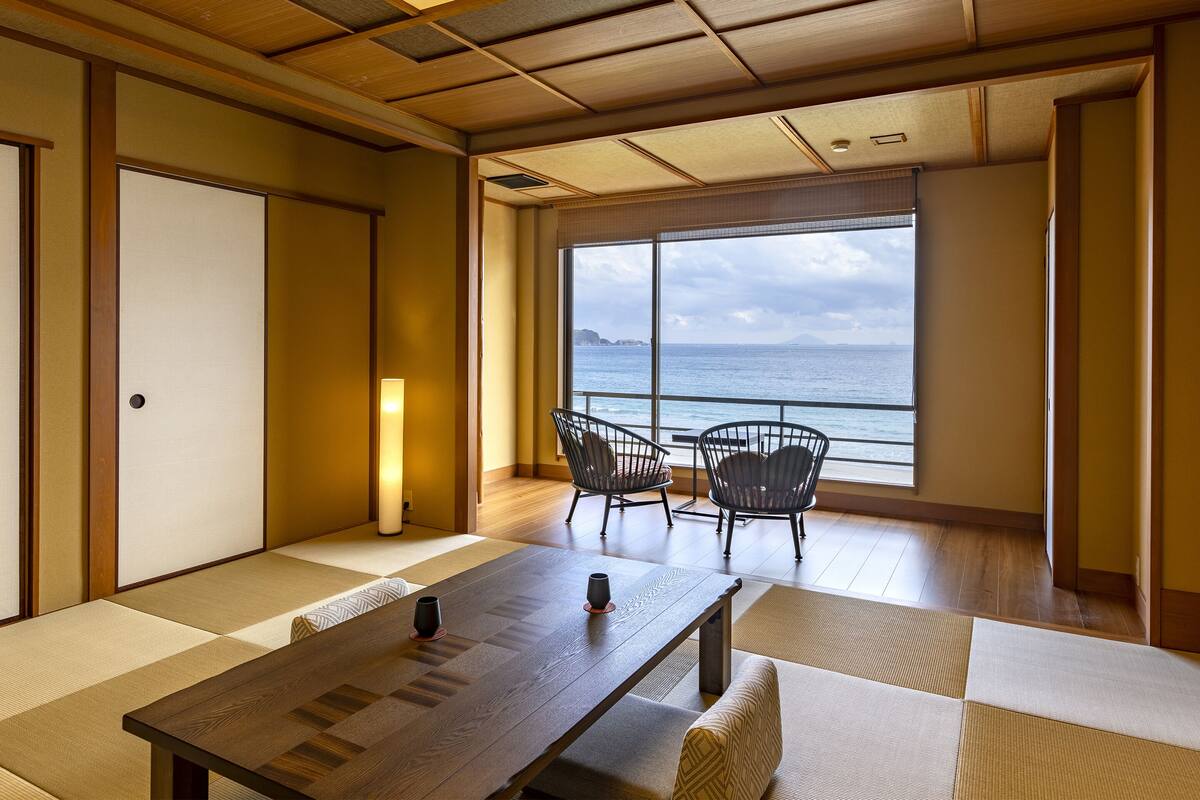 Ein traditioneller japanischer Raum mit Tatami-Böden, einem niedrigen Tisch, zwei Stühlen und einem großen Fenster mit Meerblick.