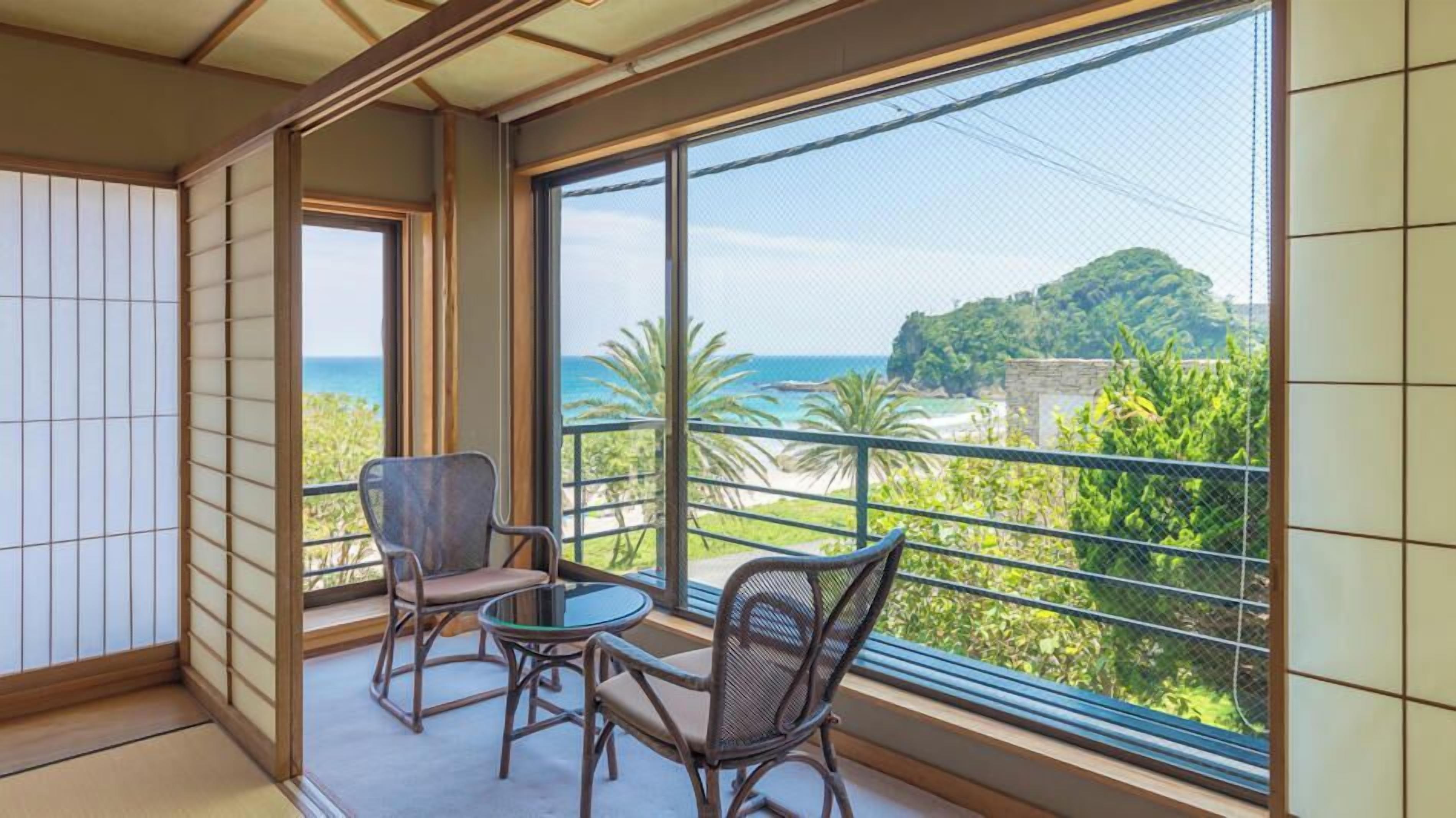 Ein Balkon mit zwei Stühlen und einem Tisch, der auf eine Küstenlandschaft mit Bäumen und Meerblick blickt.