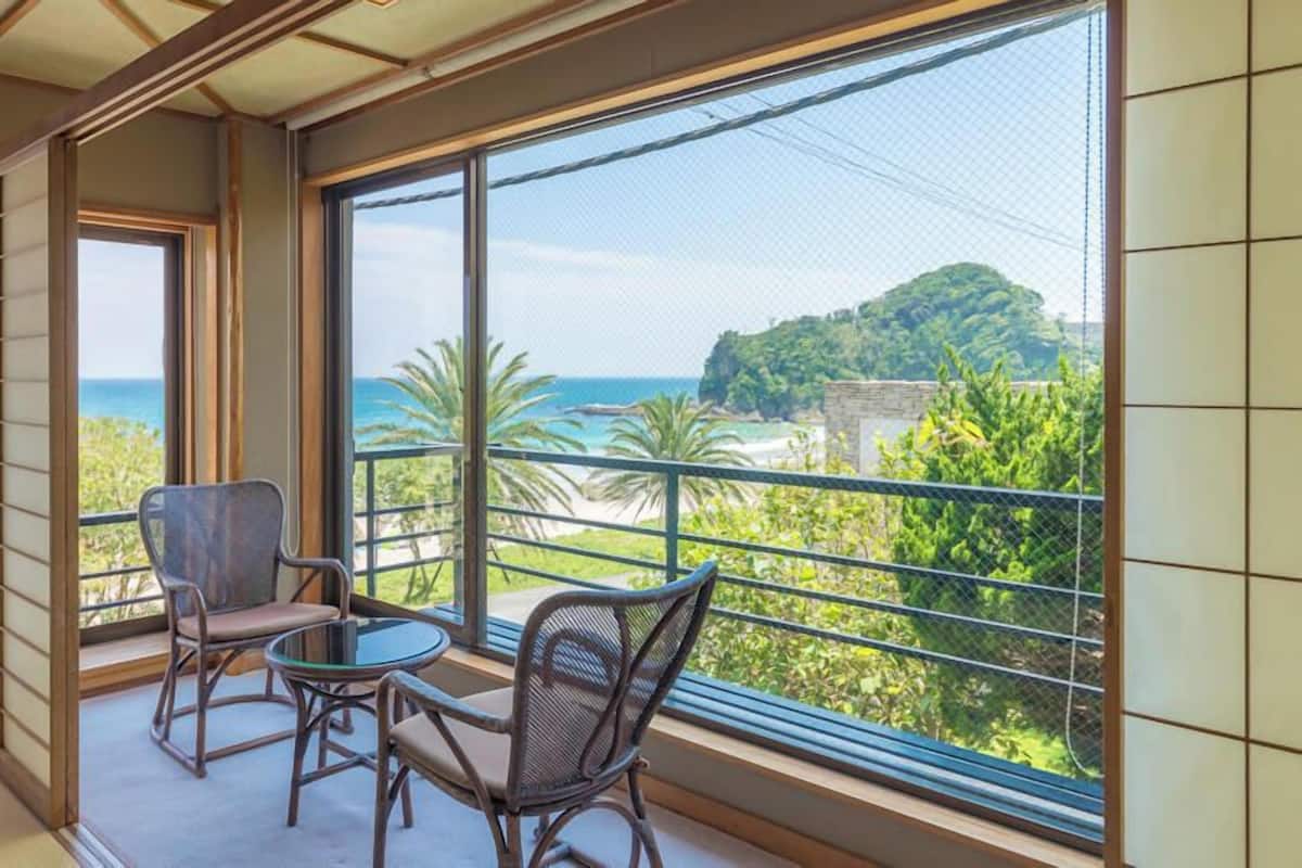 Ein Balkon mit zwei Stühlen und einem Tisch, der auf eine Küstenlandschaft mit Bäumen und Meerblick blickt.