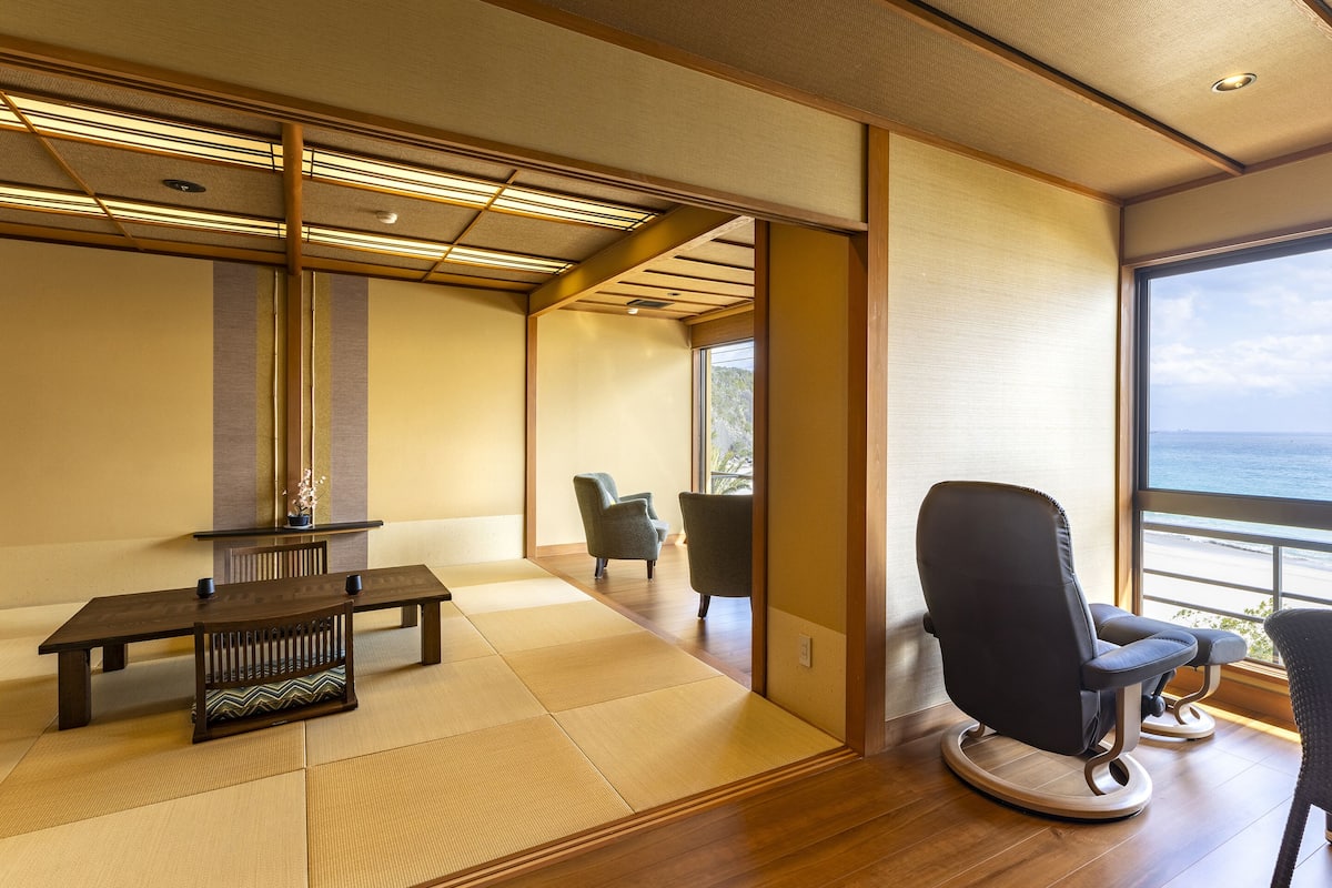 Ein Zimmer mit Tatami-Böden, einem niedrigen Tisch, zwei Stühlen und Meerblick durch ein großes Fenster.