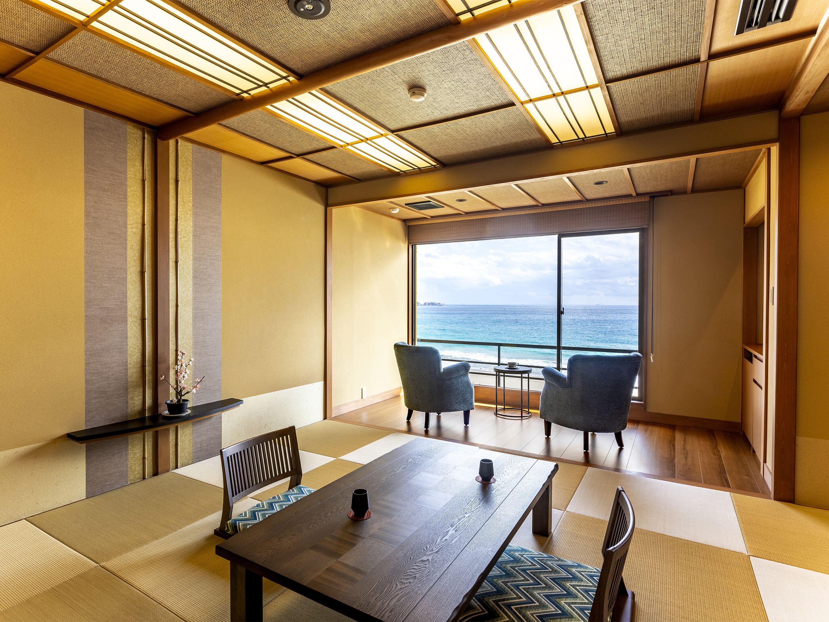 A room with a large table, two chairs, a small table with a cup, and a view of the ocean through large windows.