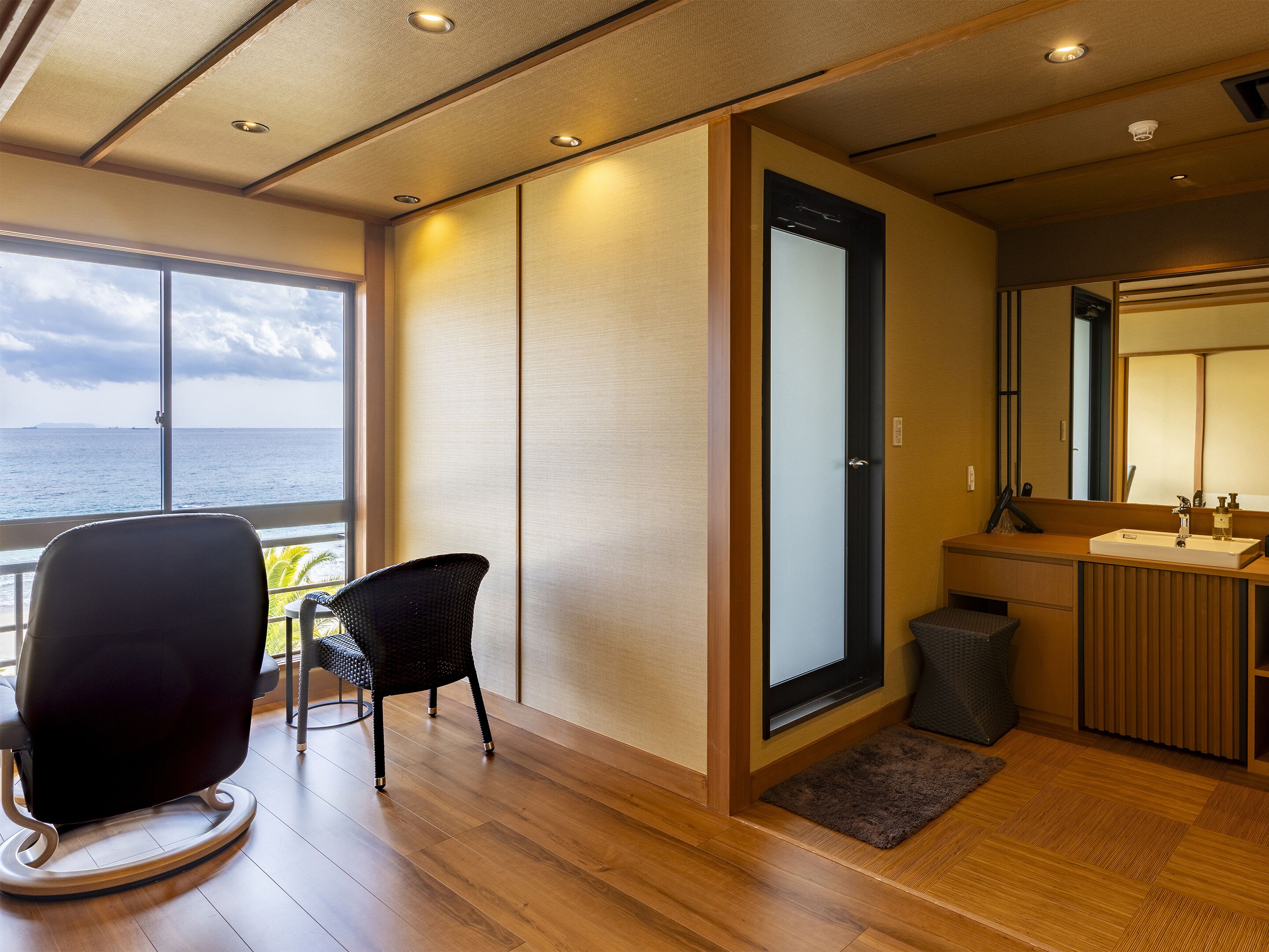 A room with a view of the sea, a black chair, a wooden floor, a small rug, and a bathroom with a sink and mirror.