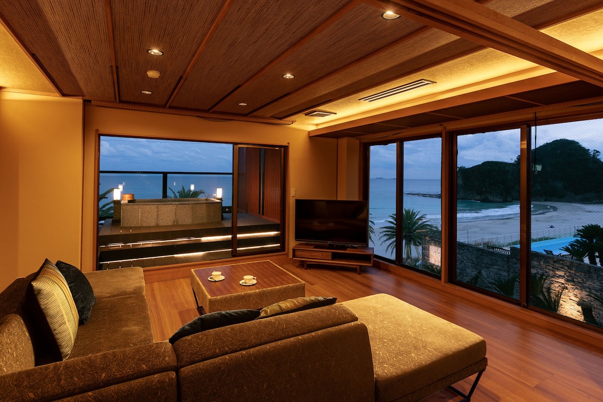 Ein großzügiger Wohnbereich mit einer großen Couch, einem Couchtisch und einem Fernseher. Es gibt einen Balkon mit Blick auf Strand und Meer.