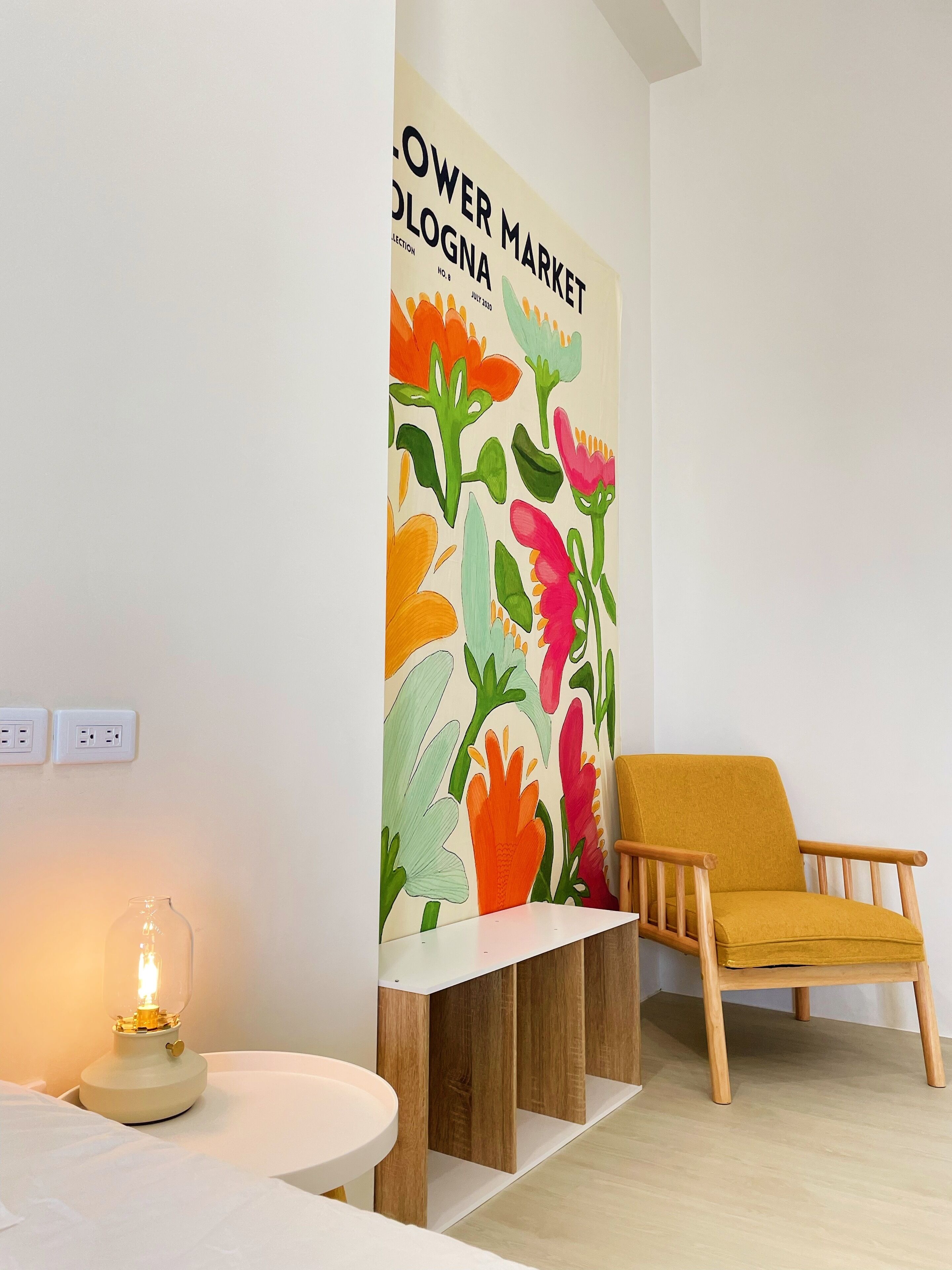 A room with a white wall featuring a colorful floral mural, a wooden chair, a table with a lamp, and a white shelf.