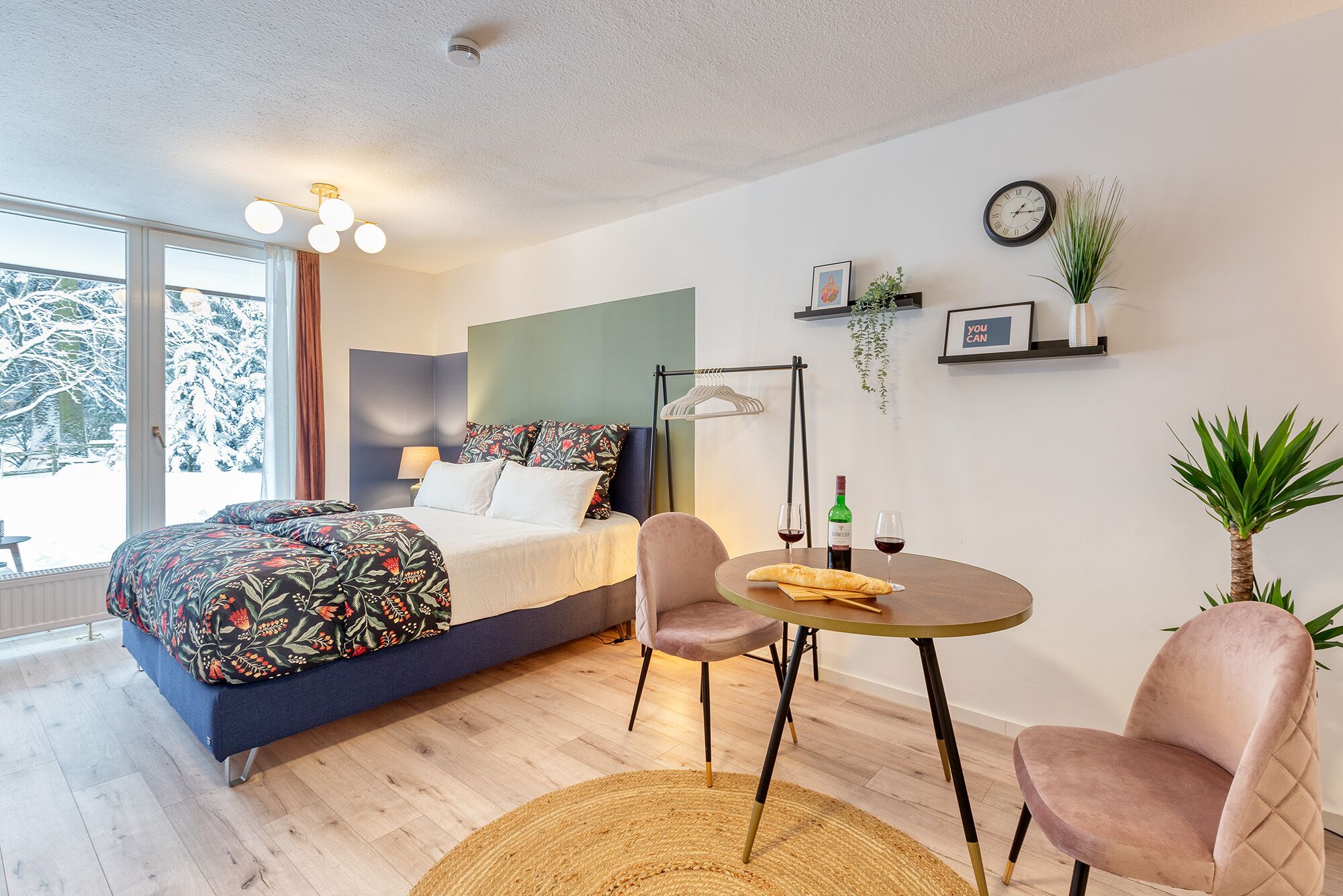A modern bedroom with a bed, bedside table, chairs, a round table, and a view of snow-covered trees through large windows.
