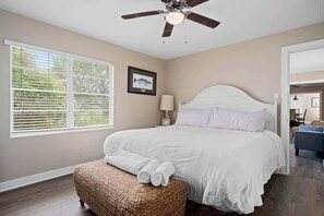 A bedroom with a bed, a ceiling fan, a window with blinds, a framed picture on the wall, and a wicker ottoman with towels.