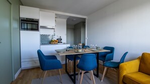 Une cuisine compacte avec des meubles blancs, un four encastré et un coin repas agrémenté de chaises bleues et d’un canapé jaune.