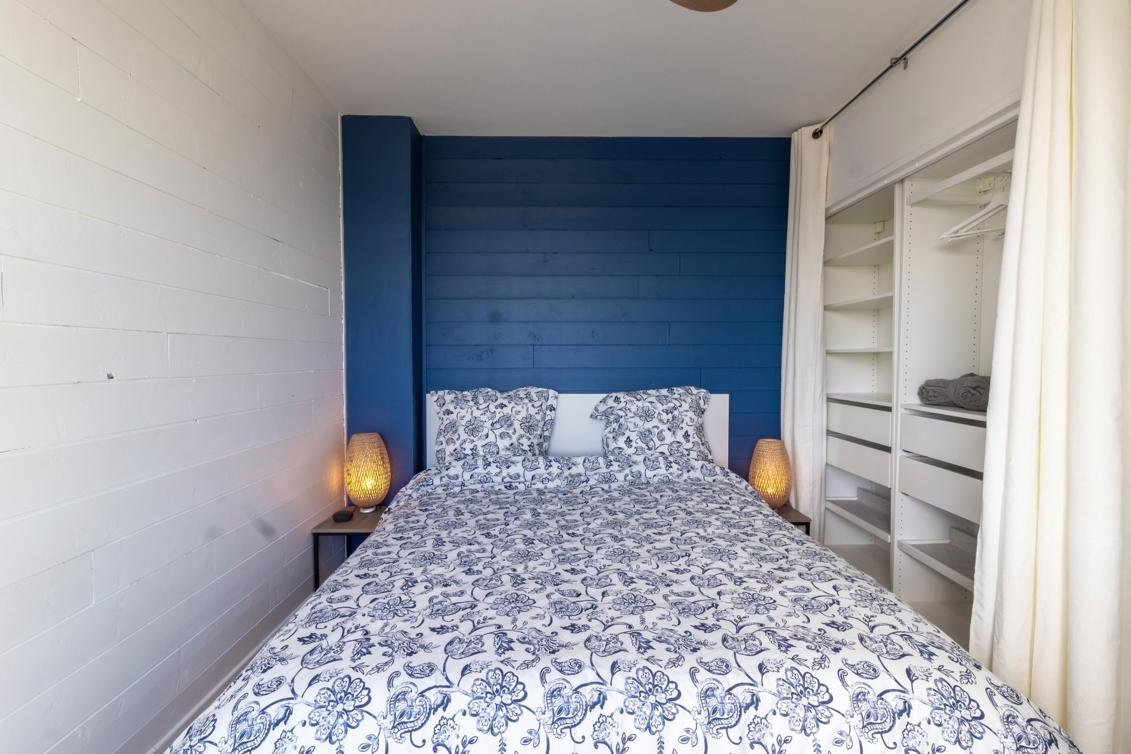 Une chambre à coucher avec une tête de lit bleue, une couvre-lit à motifs et une armoire blanche avec des étagères.