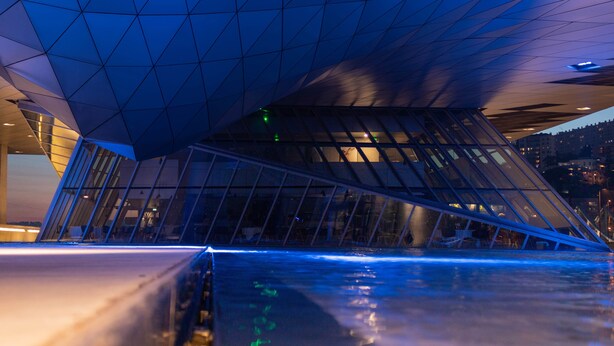 Une structure architecturale moderne au design géométrique, avec une surface d’eau réfléchissante et une vue sur le paysage urbain nocturne en arrière-plan.