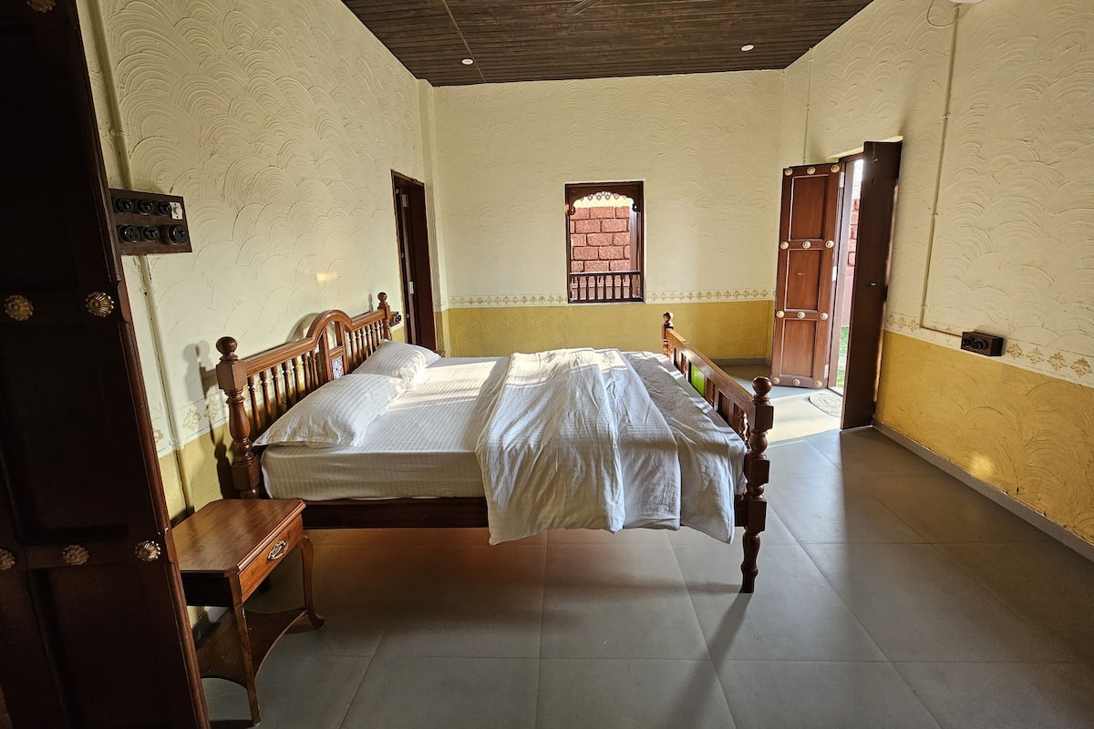 Quarto com cama, mesa de cabeceira de madeira, ventilador de teto e janela com grade decorativa.