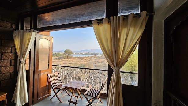 A balcony with a wooden table and chairs, curtains, and a view of a landscape with a body of water and hills. - Apla Wada (Khandala)