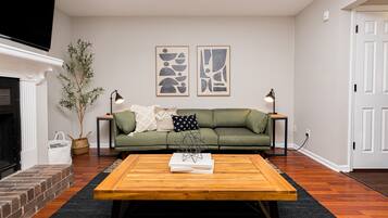 Een woonkamer met een groene bank, twee abstracte kunstwerken aan de muur, een houten salontafel, een open haard en een potplant.