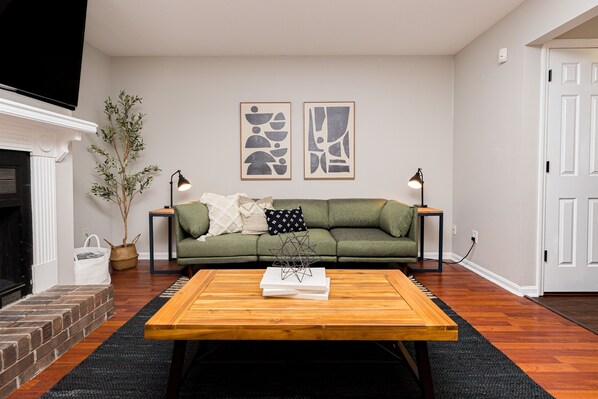 A living room with a green sofa, two abstract wall art pieces, a wooden coffee table, a fireplace, and a potted plant. - Convenient Stay | 3BR | Fenced Yard | Near RAFB (Warner Robins)