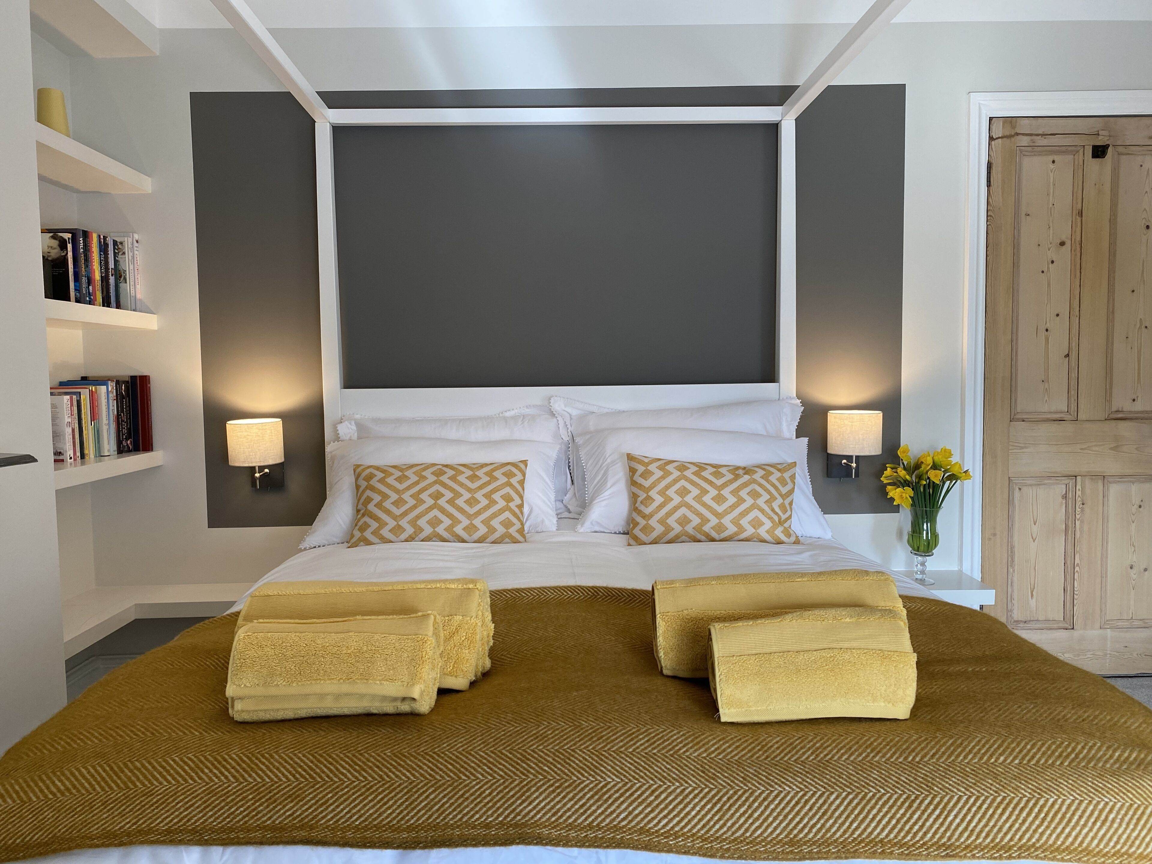 A neatly made bed with a patterned bedspread and two pillows, a bedside lamp, and a bookshelf with books.