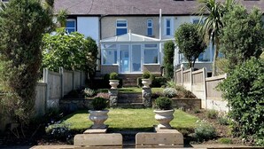 A wooden deck with two chairs, a garden with a stone pathway leading to a house with a conservatory, and a clear blue sky. - Isfyn, Boutique style cottage with stunning views in Llandudno.  (Penrhyn-side)