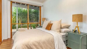 A bedroom with a large window, a bed with beige bedding, a green nightstand with a lamp, and a view of palm trees. - The Birkdale by Ready Set Stay- heated pool, WIFI, amazing and modern (Sunset Strip)